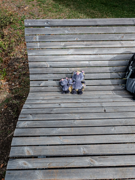 Die drei Kuscheltier-Tenreks Klein Viki, mini Christopher und Baby Viki sitzen gemeinsam auf einer hölzernen Wellenliege an der Kreuzkapelle und tanken Sonne.