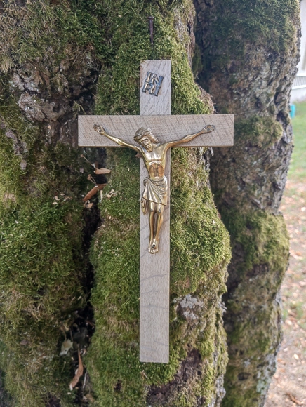 Ein kleines Holzkreuz mit metallener Christusfigur, befestigt an einer von Moos bewachsenen Baumrinde nahe der Kreuzkapelle.