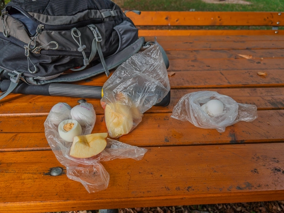 Stärkendes Wanderfrühstück auf dem Holztisch: Hartgekochte Eier und frische Apfelschnitze neben dem schwarzen Tourenrucksack.
